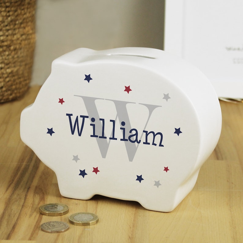 Może przedstawiać: Biała ceramiczna skarbonka z szarym literą W i imieniem "William" w granatowych literach. Skarbonka jest ozdobiona czerwonymi, niebieskimi i szarymi gwiazdami.