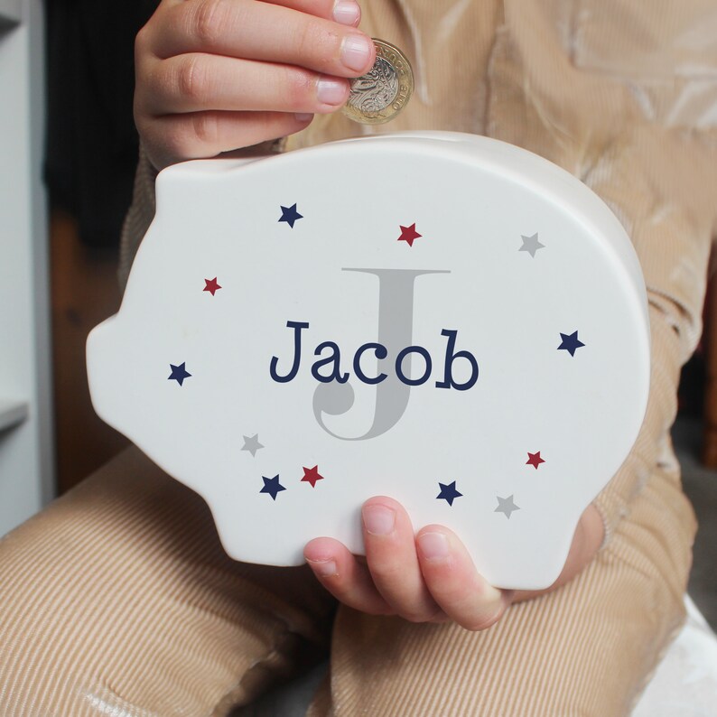 Może przedstawiać: Biała ceramiczna skarbonka z szarym literą J i imieniem Jacob w niebieskich literach. Skarbonka jest ozdobiona czerwonymi, niebieskimi i szarymi gwiazdami.
