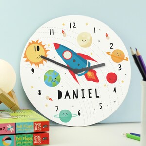 May include: A white round clock with a rocket, planets, and stars. The clock face has black numbers and hands. The name "DANIEL" is printed below the rocket.