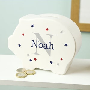 Może przedstawiać: Biała ceramiczna skarbonka z niebieską literą 'N' i imieniem 'Noah' w niebieskich literach. Skarbonka jest ozdobiona czerwonymi i szarymi gwiazdami.