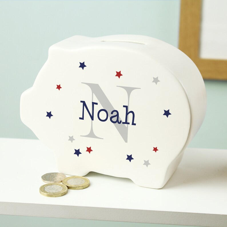 Może przedstawiać: Biała ceramiczna skarbonka z niebieską literą 'N' i imieniem 'Noah' w niebieskich literach. Skarbonka jest ozdobiona czerwonymi i szarymi gwiazdami.