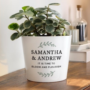May include: A white ceramic plant pot containing a variegated green and white plant. The pot features a sage green leaf graphic above and below the black text "SAMANTHA & ANDREW" and "IT IS TIME TO BLOOM AND FLOURISH" printed on the front surface.