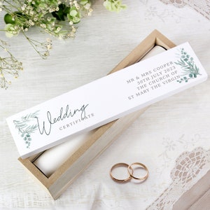 May include: A white box with a wooden lid and a white label that says "Wedding Certificate" with green foliage and the text "MR & MRS COOPER 30TH JULY 2023 THE CHURCH OF ST MARY THE VIRGIN".