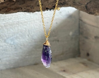 May include: A gold chain necklace with a purple amethyst crystal pendant. The crystal is wrapped in gold wire and has a natural, raw shape.