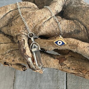May include: Two silver chains with pendants. One pendant is a clear quartz crystal point, the other is a gold evil eye charm with a blue eye.