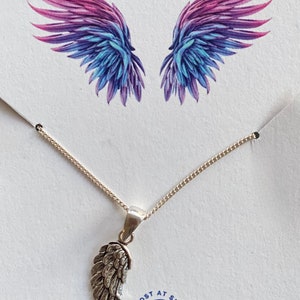 May include: A silver necklace with a single angel wing pendant. The necklace is on a white background with a pair of watercolor wings in the background. The text "Lost at Sea. You & Me" is printed below the necklace.