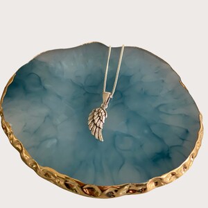 May include: A silver angel wing pendant necklace on a silver chain, displayed on a blue and gold marbled surface.