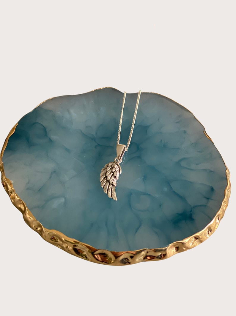 May include: A silver angel wing pendant necklace on a silver chain, displayed on a blue and gold marbled surface.
