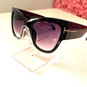 May include: Black cat eye sunglasses with dark purple gradient lenses. The sunglasses have a gold accent on the temple arms and are displayed on a clear acrylic stand.  These fashionable sunglasses feature a sleek, modern design.