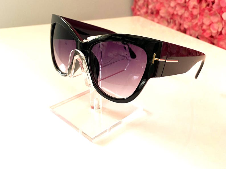 May include: Black cat eye sunglasses with dark purple gradient lenses. The sunglasses have a gold accent on the temple arms and are displayed on a clear acrylic stand.  These fashionable sunglasses feature a sleek, modern design.