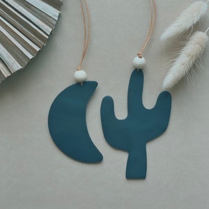 Puede incluir: Dos adornos de cerámica azul oscuro, una luna creciente y un cactus, colgando de cordones marrones con cuentas blancas.