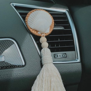 Pode incluir: Uma concha branca com uma moldura de vime tecida pendura de uma borla branca. A borla está presa a uma conta de madeira e uma conta de madeira menor. A concha e a borla estão penduradas na ventilação de um carro.