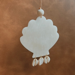 Könnte beinhalten: Ein weißer Keramik-Muschel-Anhänger mit einer Schnur zum Aufhängen. Die Muschel hat eine strukturierte Oberfläche und ist mit kleinen weißen Muscheln verziert, die an der Unterseite hängen.