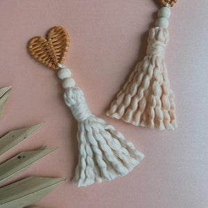May include: Two woven heart shaped ornaments with tassels. The hearts are made of natural wicker and the tassels are made of cream and peach coloured yarn.