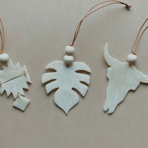 May include: Four white ceramic ornaments with brown leather cords. The ornaments are shaped like a monstera leaf, a bull skull, a geometric design, and a square.