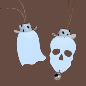 May include: Two white Halloween ornaments: a ghost and a skull, each wearing a black and white spotted cowboy hat. The skull ornament has a small silver disco ball attached. Both ornaments hang from brown cords against a brown backdrop.