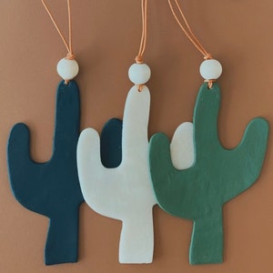 Puede incluir: Tres adornos de cactus de madera colgando de cordones marrones. Las formas de cactus están pintadas de negro, blanco y verde.