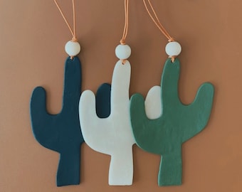 Peut inclure: Trois ornements en bois en forme de cactus suspendus à des cordons bruns. Les formes de cactus sont peintes en noir, blanc et vert.