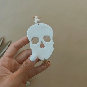 Puede incluir: Adorno de calavera de cerámica blanca con un amuleto de sol dorado. La calavera tiene un diseño sencillo con dos agujeros para los ojos y una nariz.