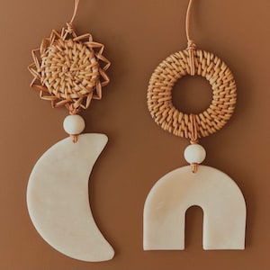 May include: Two hanging decorations, one a white crescent moon and the other a white arch, both with a woven rattan sun and circle above them.