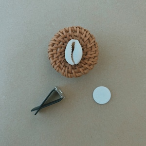 Könnte beinhalten: Ein gewebter Rattan-Untersetzer mit weißem Muschelakzent. Daneben befinden sich ein schwarzer Auto-Lüftungsclip und ein kleines weißes rundes Pad. Auf neutralem Hintergrund.