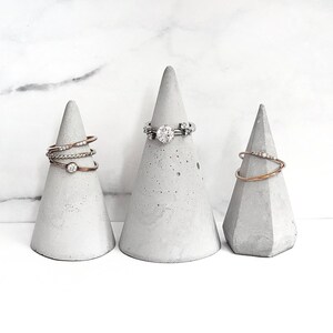 May include: Three grey concrete ring holders shaped like cones. The cone in the centre has a silver ring with a clear stone on it. The cones on either side have a silver ring and a gold ring with clear stones on them.