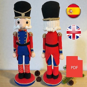 Puede incluir: Muñeco cascanueces de ganchillo con uniforme de soldado. El muñeco tiene un sombrero negro, bigote blanco y uniforme rojo, azul y dorado. El muñeco está sobre una base roja y azul. La imagen muestra el frente y la parte posterior del muñeco.