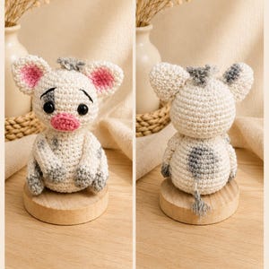 May include: A handmade crochet cow doll with a white body, gray spots, and pink inner ears. The doll has black eyes and a pink mouth. It is displayed on a small wooden stand. The cow has a gray tail.