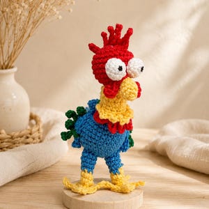 May include: A handmade crochet rooster figurine. The rooster has a red comb, white eyes, and a yellow beak. Its body is blue with yellow feet and a yellow and red collar. The figurine is standing on a small wooden base.