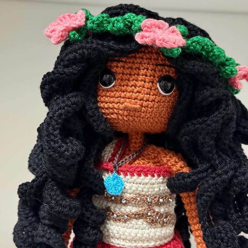 May include: A handmade crochet doll with black curly hair, brown skin, and large black eyes. The doll wears a floral crown with pink flowers and a white and brown top with a blue pendant necklace. The doll's dress is red.