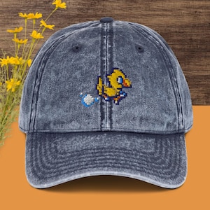 May include: A denim blue baseball cap with a pixelated yellow bird design. The cap has a curved brim and a distressed, vintage look. The bird is running with a blue and white trail behind it.