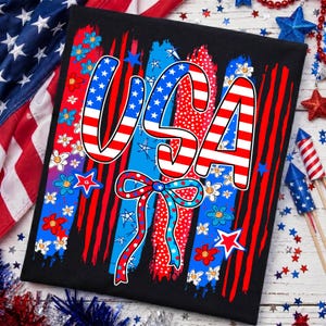 Peut inclure: T-shirt noir avec un motif patriotique "USA". Les lettres sont rayées rouge et blanc avec des détails d'étoiles bleues. Le motif comprend des coups de pinceau rouges et bleus, des fleurs et un nœud.