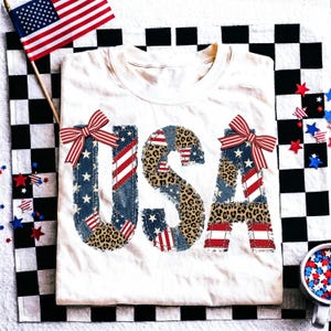 Peut inclure: T-shirt crème avec le mot "USA" dans un motif patchwork. Les lettres présentent des motifs denim, léopard et drapeau américain, avec des nœuds rayés rouges et blancs. Un drapeau américain et des confettis patriotiques complètent le look.