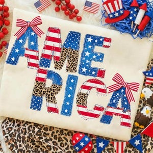 Op de afbeelding: Crèmekleurig T-shirt met het woord "AMERICA" in patriottische kleuren en luipaardprint. De letters zijn versierd met rood, wit en blauwe patronen, en het ontwerp bevat strikken en sterren. Extra patriottische decoraties omringen het shirt.