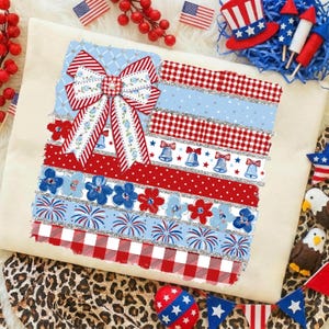 Peut inclure: Un coussin décoratif sur le thème patriotique, avec un motif rappelant le drapeau américain. Il présente un grand nœud, des motifs floraux et de cloches, ainsi que des motifs rouges, blancs et bleus. Des décorations patriotiques supplémentaires entourent le coussin.