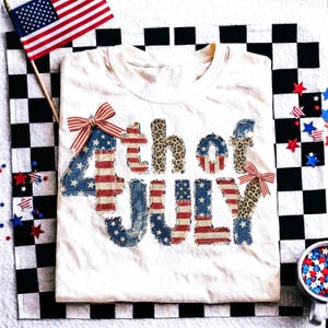 Peut inclure: Un t-shirt crème avec le texte "4th of July" dans un design patriotique. Les lettres présentent des motifs de drapeau américain et un imprimé léopard. Le t-shirt est orné de nœuds rouges, blancs et bleus et est posé sur une surface à carreaux noirs et blancs.