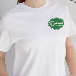 Pode incluir: Camiseta branca com um logotipo circular verde que diz "Merlotte's Bar and Grill".