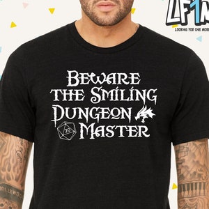 Op de afbeelding: Zwart T-shirt met witte tekst die "Beware the Smiling Dungeon Master" luidt. Een witte 20-zijdige dobbelsteen en een draak zijn ook in het ontwerp opgenomen.