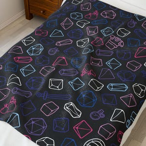 Pode incluir: Um cobertor preto com um padrão repetido de formas geométricas e ilustrações de fantasia brancas, rosa, azuis e roxas. O cobertor está em uma cama com uma almofada branca. O cobertor tem o texto "LF1M".