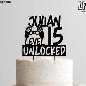 May include: A white frosted cake with a black cake topper that reads "JULIAN 15 LEVEL UNLOCKED" with a controller graphic. The cake sits on a wooden cake stand.