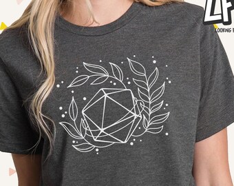 Puede incluir: Una camiseta gris oscuro con un gráfico blanco de un dado geométrico rodeado de hojas. El gráfico está centrado en la camiseta.