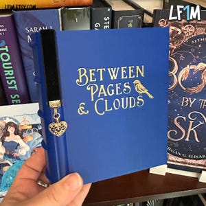 Puede incluir: Un libro azul con letras doradas que dice "Between Pages & Clouds" y una ilustración de un pájaro. Un marcapáginas de terciopelo negro con un dije de corazón dorado cuelga del libro. El libro se exhibe entre otros libros en un estante.