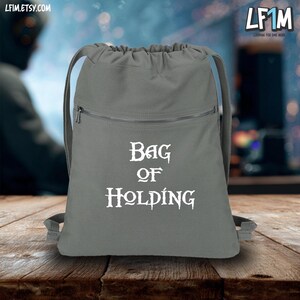 May include: Gray drawstring backpack with a zippered front pocket. The words "Bag of Holding" are printed in white. The bag has a drawstring closure and shoulder straps. The Etsy shop LF1M is in the top right corner.