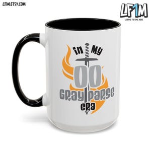 May include: White ceramic mug with a black handle and rim. The mug has a graphic design of a sword with flames and the text "in my 00 gray parse era".