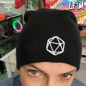 May include: A black knit beanie featuring a white geometric design on the front. The beanie is made of a ribbed knit material and is worn low on the head. The text "LF1M" is visible in the background.