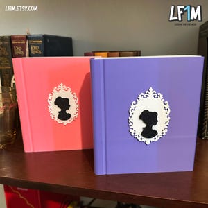 May include: Two hardcover books, one pink and one purple, with a decorative silhouette of a woman in a white ornate frame. The books are on a wooden shelf with other books in the background. The website LF1M.ETSY.COM is visible.