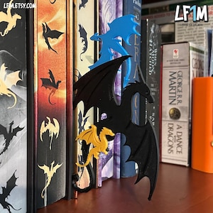 May include: Dragon-shaped bookmarks in black, blue, and gold are inserted into the pages of various books. The bookmarks are made of a solid material and have detailed wing and body designs. The books are on a shelf, with the text "LF1M" visible.