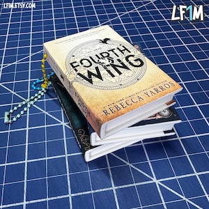 May include: A stack of miniature books, including one titled "Fourth Wing" by Rebecca Yarros. The top book has a tan cover with a circular design and the title in bold letters. A beaded chain with blue and yellow beads is visible.