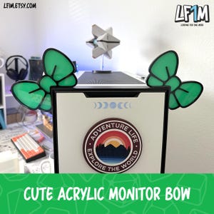 May include: Green acrylic monitor bows with black outlines, shaped like butterfly bows, are displayed on a white computer monitor. The monitor has a circular sticker that says "Adventure Life Explore the World." The text "CUTE ACRYLIC MONITOR BOW" is at the bottom.