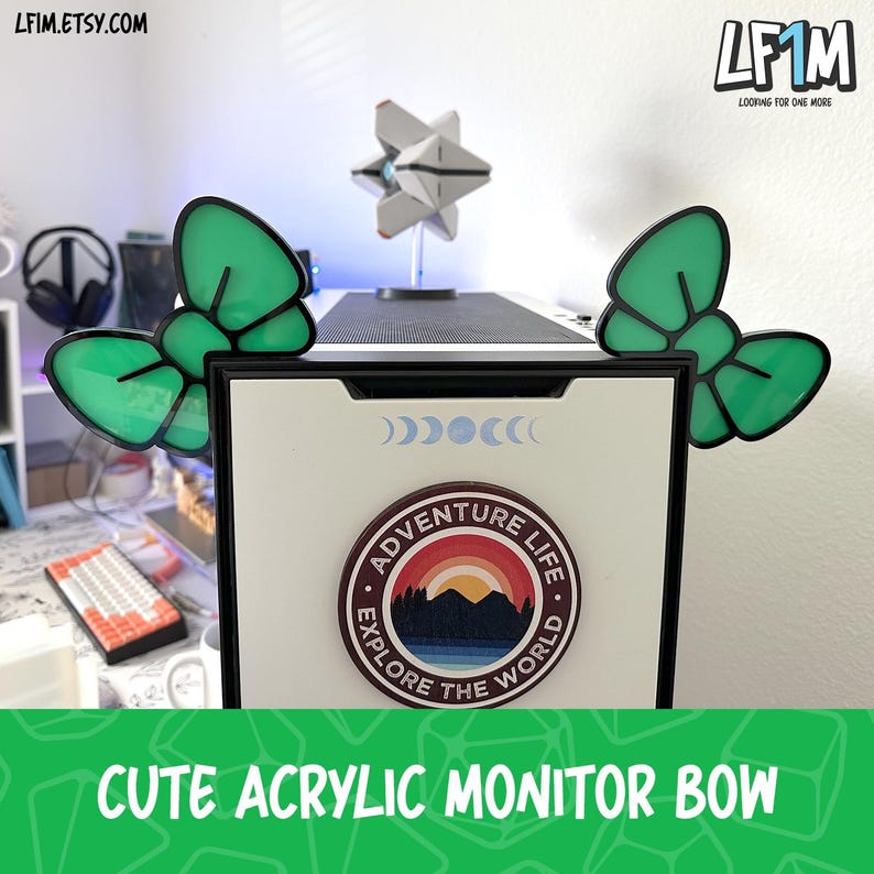 May include: Green acrylic monitor bows with black outlines, shaped like butterfly bows, are displayed on a white computer monitor. The monitor has a circular sticker that says "Adventure Life Explore the World." The text "CUTE ACRYLIC MONITOR BOW" is at the bottom.